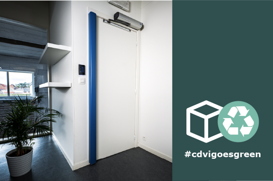 En vit dörr med ett blått vertikalt handtag och en elektronisk kodpanel bredvid dörrkarmen. Rummet har en modern design med en krukväxt i förgrunden och hyllor på väggen. Till höger finns en grön banner med en återvinningssymbol och texten #cdvigoesgreen.